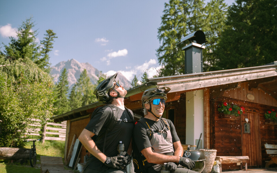 Mountainbiker ruhen sich an einer Hütte in den Dolomiten, Italien, aus, tragen Helme und Sonnenbrillen und genießen den sonnigen Tag.