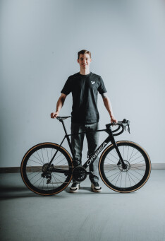 Lächelnder Radfahrer posiert mit einem Orbea-Rennrad vor einem schlichten Hintergrund für eine Werbeaktion für Fahrradausrüstung.