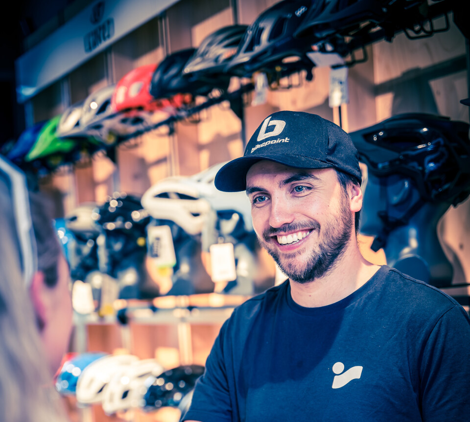 Ein lächelnder Mitarbeiter eines Fahrradgeschäfts übergibt einem Kunden einen Kauf; Fahrradhelme werden im Hintergrund ausgestellt.