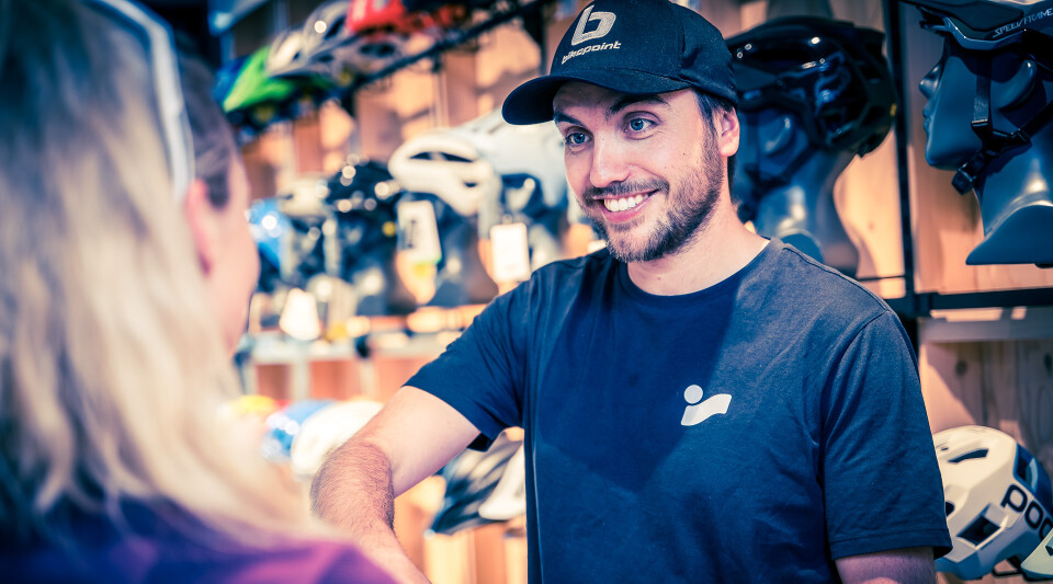 Freundlicher Fahrradladen-Mitarbeiter hilft einem Kunden beim Kauf von Fahrradbekleidung. Helme werden ausgestellt.