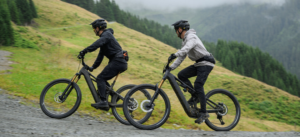 Zwei Mountainbiker fahren mit Elektrofahrrädern bergauf auf einem Schotterweg in einer grünen Berglandschaft.