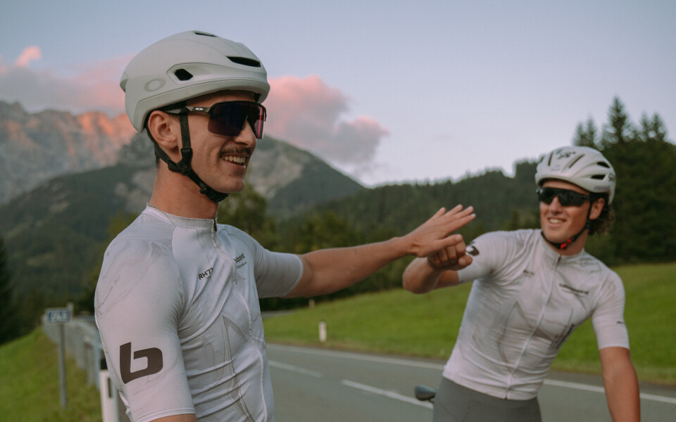 Zwei Radfahrer geben sich auf einer Straße vor einer Bergkulisse ein High-Five, sie tragen Radsportbekleidung und Helme.