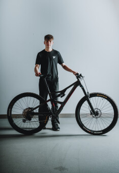 Junger Mann posiert mit einem schwarzen vollgefederten Mountainbike in einer Studio-Umgebung.