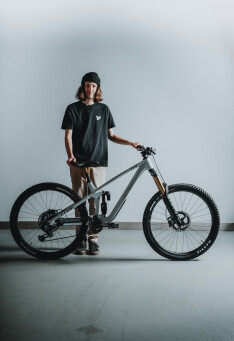 Junger Mann mit langen Haaren posiert mit einem grauen vollgefederten Mountainbike in einer Studio-Umgebung.